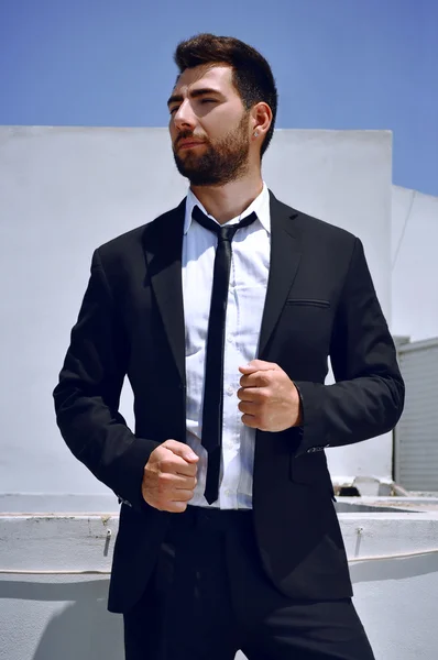 Attractive Spanish looking young man with a beard and dark hair and eyes is wearing a black suit and tie with a white shirt.