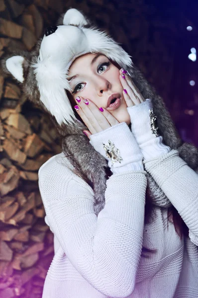 Teenage girl with red hair and green eyes wearing a furry husky hat with ears and pompoms, and knitted hand warmers with diamond detail is touching her cold cheeks with a surprised expression on her face.