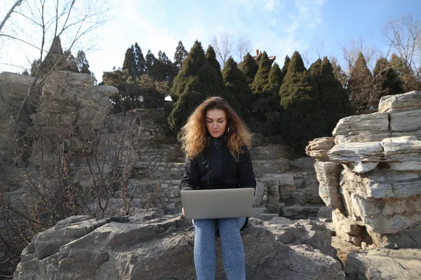 Beautiful young girl sitting with a laptop in the park on a background of mountains. The possibilities are endless on the Internet to work, learn, communicate.