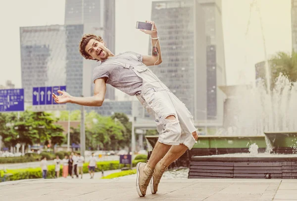 Young stylish man is dancing in the street with headphones and holding a mobile phone. Guy in motion on tiptoes. Guy having fun and smiling.