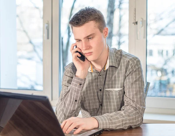 Young man calls on the phone sitting at home on the table in front of the laptop.