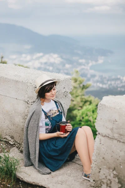 Young fashion woman sitting on a top of the mountain drinking tea