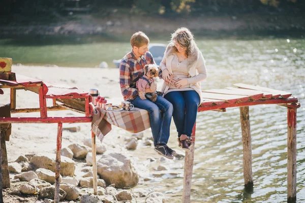 Young future parents and their dog in a funny costume sitting on a wooden bridge and having picnic near lake