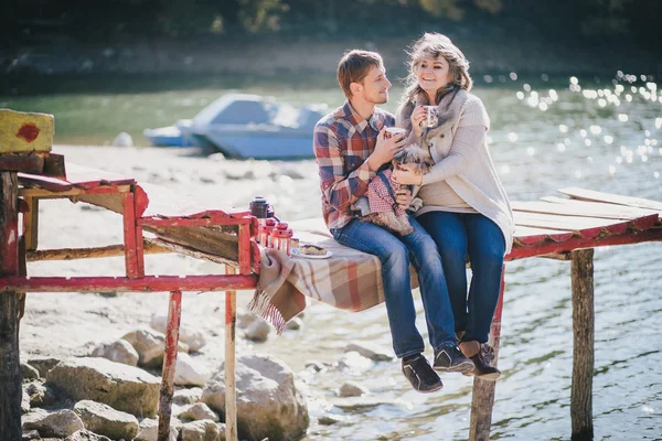 Young future parents and their dog in a funny costume sitting on a wooden bridge and having picnic near lake