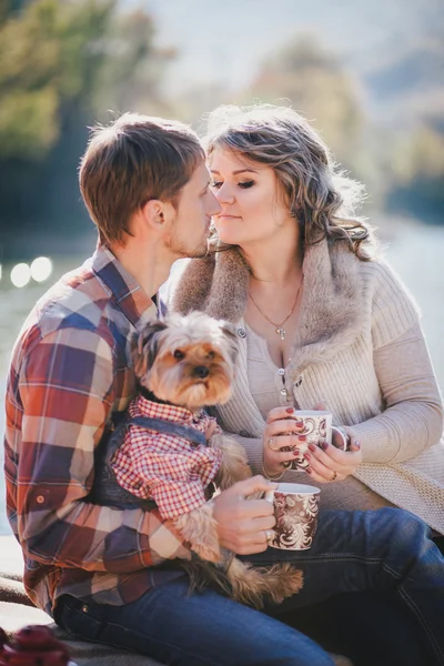 Young future parents and their dog in a funny costume sitting on a wooden bridge and having picnic near lake