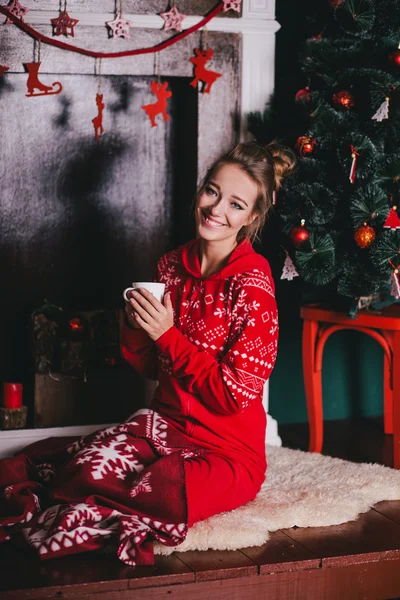 Young beautiful woman in a red warm pajamas with scandinavian ornaments sitting near decorative fireplace and drinking hot tea or cocoa