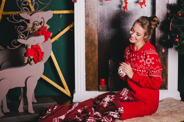 Young beautiful woman in a red warm pajamas with scandinavian ornaments sitting near decorative fireplace and drinking hot tea or cocoa