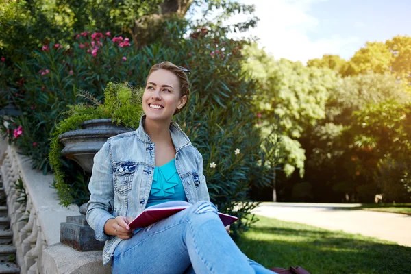 Young cheerful Caucasian female is smiling to someone while is relaxing outdoors with book