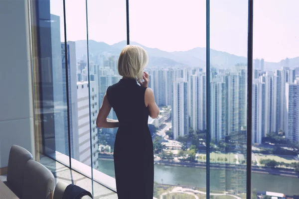 Female CEO is calling via cell telephone, while is standing in office interior near window with cityscape view and copy space