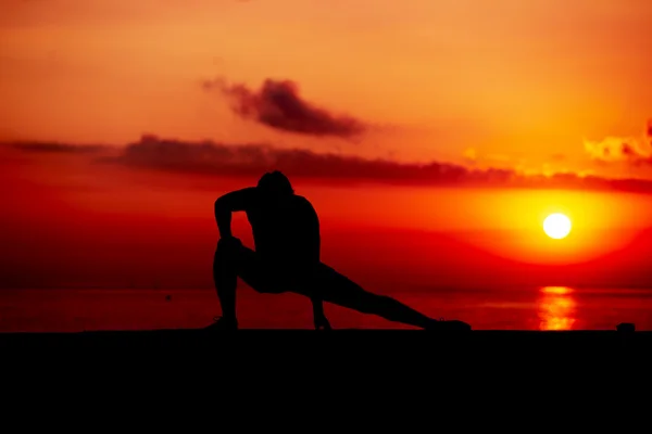 Silhouette of young man at cross-training during colorful sunrise on the beach, athletic runner with muscular body doing stretching legs exercise outdoors, fitness and healthy lifestyle concept