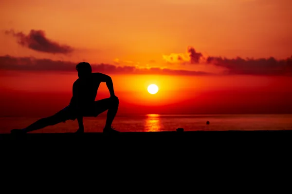Silhouette of young man at cross-training during colorful sunrise on the beach, athletic runner with muscular body doing stretching legs exercise outdoors, fitness and healthy lifestyle concept
