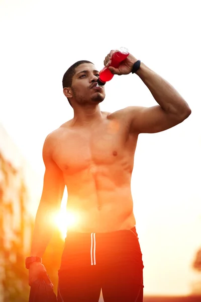 Portrait of athletic build dark skinned man with muscular body refreshing with energy drink after run at sunset outdoors