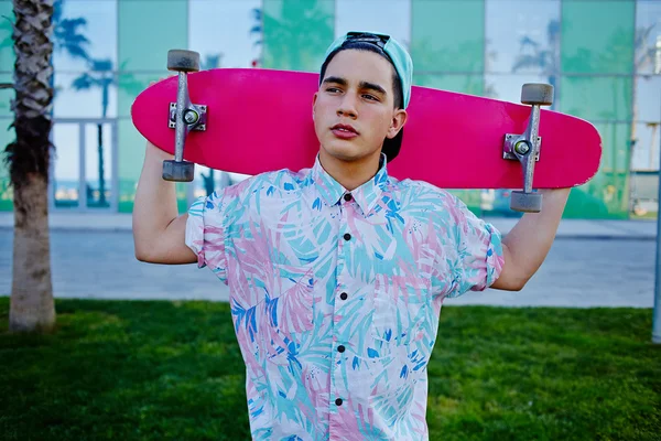 Young stylish man in bright summer clothes standing with pink long-board on beautiful glass background with reflected palm trees, cool teenager holding his long board looking away prank