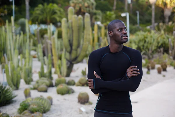 Attractive athlete resting after workout standing in beautiful cactus park at evening, fit man in black active clothes taking break after run outdoors, black man with crossed hands looking away