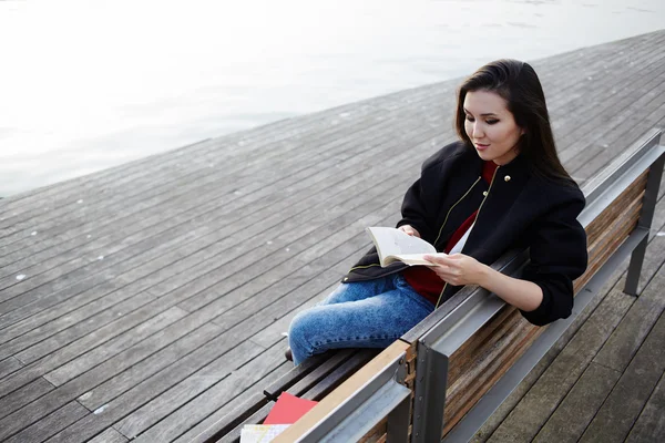 Asian student girl reading book seated on the bench, attractive college student reading book during her class break outdoors, young woman reading book on the wooden bench