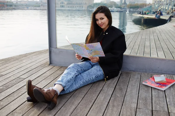 Attractive tourist girl resting after walk sitting on wooden pier in sea port, beautiful student girl holding city map in hands smiling, charming asian girl sitting with open city map on wooden pier