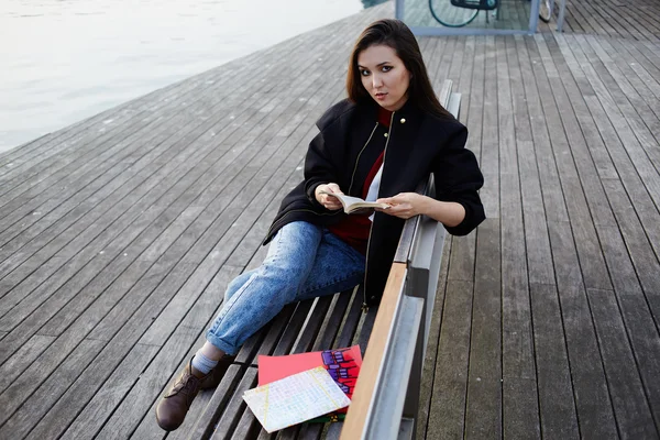 Beautiful girl holding an open book sitting on wooden bench near artificial lake, tourist woman resting after walk in new city reading some book, attractive asian girl at leisure time outdoors