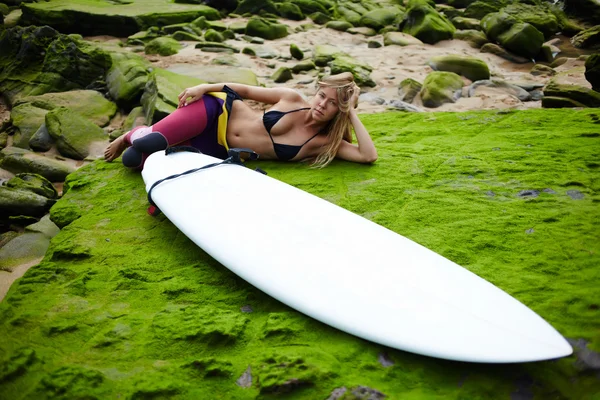 Beautiful blond girl in swimsuit relaxing after surfing lying on the beach with copy space surf board
