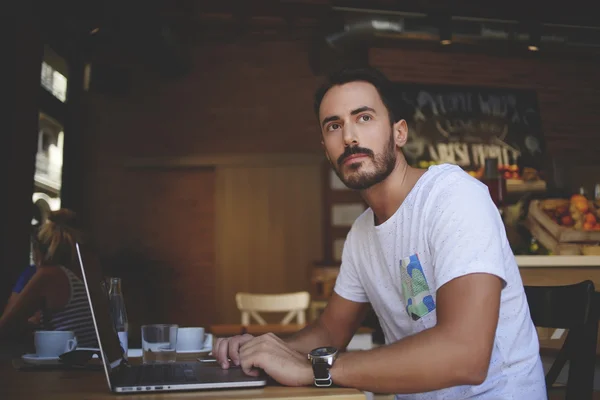 Male developer comes up with ideas for website while sitting front open laptop computer in cozy restaurant interior, young man thinking about something during work on net-book in modern coffee shop