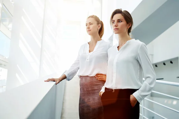 Half length portrait of a two female managing directors resting in hallway after successful presentation, young businesswomen concentrated looking in window while standing in modern office interior