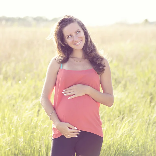 Young beautiful pregnant woman holding tummy smiling, in red a light summer dress, happy on meadow the grass