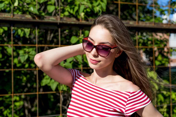 Beautiful girl in a bright red T-shirt, fashion lifestyle, posing against the backdrop of fence with green leaves