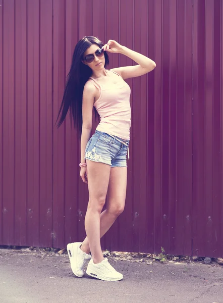 Beautiful girl with glasses shorts and pink blouse stands in front of a wooden fence, stylish fashion Sneakers.