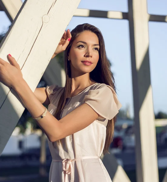 Portrait of a beautiful girl in  dress standing at the wooden pillars, resting after work, fashion style, urban life, in  summer park on  bright sunny day. Tanned brunette business.