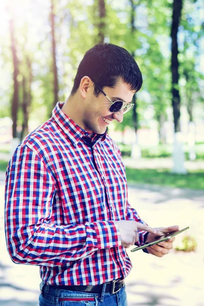 The man in the summer in the park, reading glasses, looking a map and news feed on social networks, happy smiles watching  video film in jeans bright sunny day. Idea concept businessman city plan