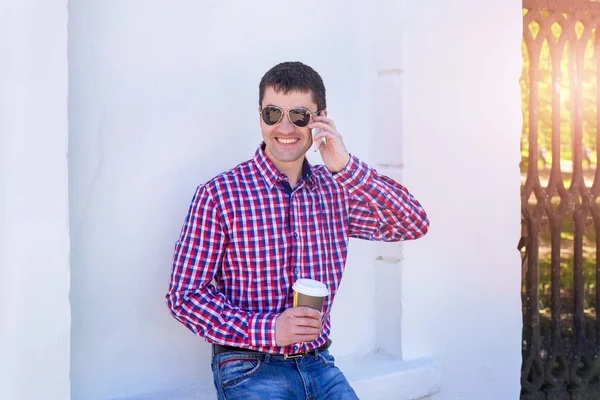 Man in  shirt and jeans  glasses, talking on the phone,  smartphone, the concept of summer,  businessman  vacation. City lifestyle.  the street in the park. Happy smiling, emotional. In his hand  mug