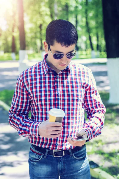 Man in  shirt and jeans  sunglasses, holding  mobile phone coffee or tea, read the conversation messages on your , the concept of summer,  businessman  vacation. City lifestyle.  the street in the