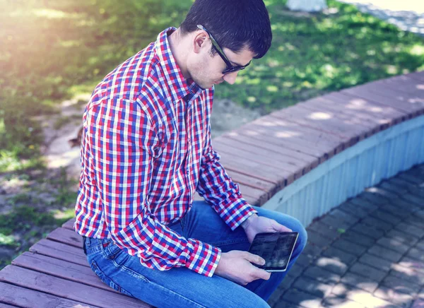 Man in  shirt and jeans  sunglasses, video looks on the tablet corresponds to the social networks, in the summer  the bench, the concept of  businessman  vacation. City lifestyle.  the street in the