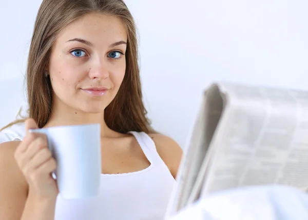 A pretty young woman reading the newspaper in bed and enjoying a cup of tea