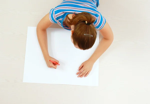 Top view of cute young woman thinking with a pen in her mouth while lying on floor and drawing.