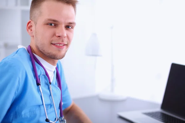 Portrait of a smiling male doctor with laptop sitting at desk in medical office