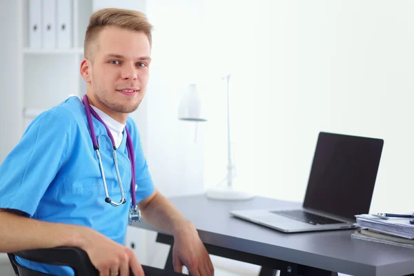 Portrait of a smiling male doctor with laptop sitting at desk in medical office