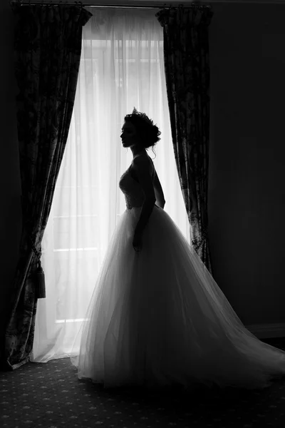 Beautiful delicate sexy bride happy woman with a crown on her head by the window with a large wedding bouquet in a luxurious white dress