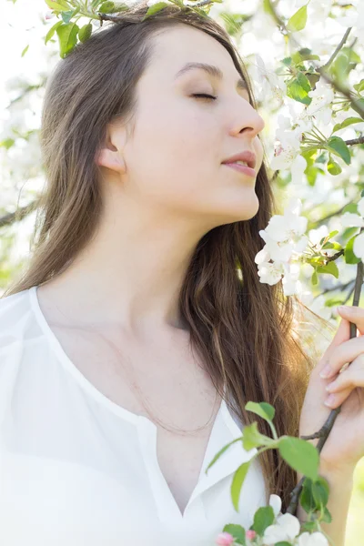 Beautiful sweet girl in a white shirt with blooming apple trees in the garden walks bright warm summer day