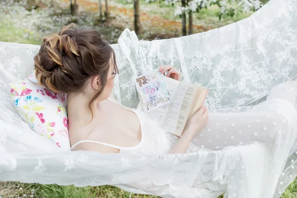 Beautiful sexy cute girl in a light white dress in apple blossoming garden sees on the hammock with a book