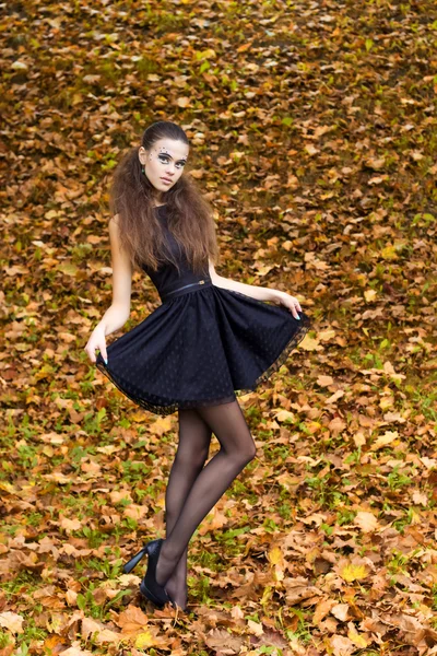 Beautiful young girl on the background of the leaves in autumn day on the street with fantasy makeup in a black dress with big sexy lips