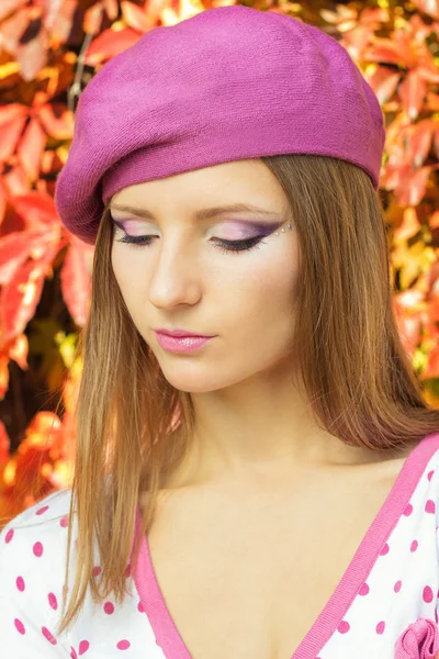 Beautiful sexy girl in a pink hat with beautiful makeup in the white jacket in pink polka dot stands on a Sunny autumn day in the Park near red leaves