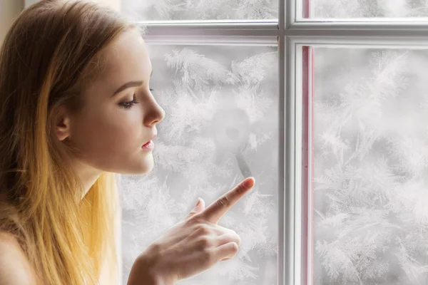 Beautiful young girl sitting by the window winter day and draws the sun on the frozen window