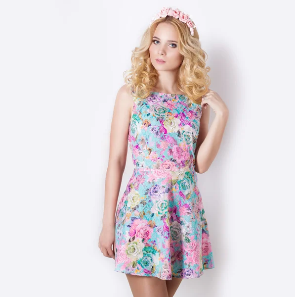 Beautiful sexy modest sweet tender girl with blonde curly hair with a wreath of flowers in their hair standing on a white background in a blue summer dress