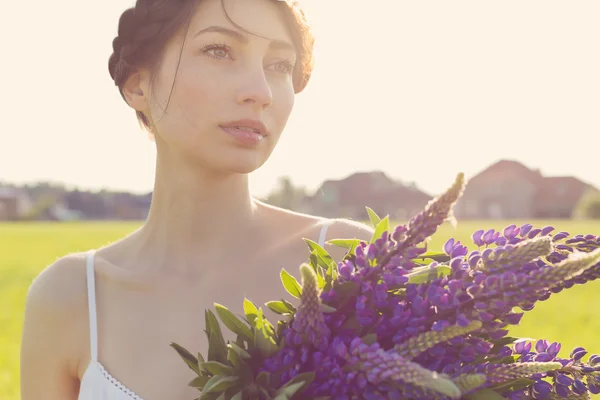 Beautiful sexy girl with a sweet lupine flowers with beautiful hair in a white sundress standing in a field at sunset in the warm summer sun