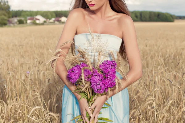 Beautiful sexy slim girl in a blue dress in the field with a bouquet of flowers and ears of corn in his hands at sunset on a sunny day