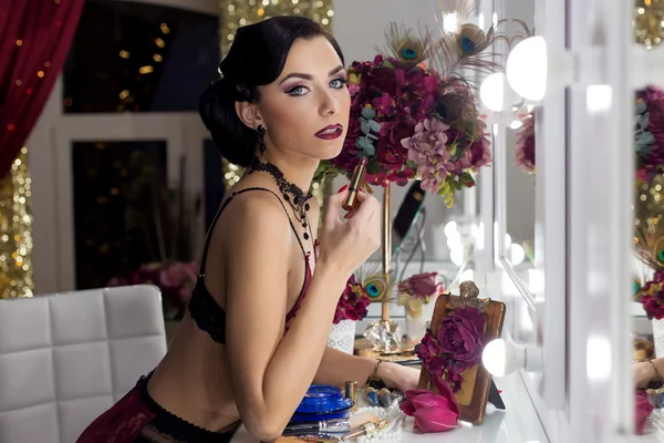 Beautiful sexy young woman applies lipstick before a mirror in the dressing room in lacy underwear in a retro style with bright make-up and beautiful evening hairstyles , shot in the style of the 20s