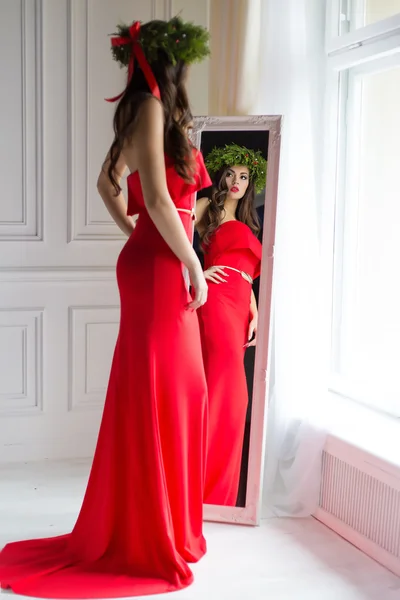 Beautiful sexy woman in elegant long evening red dress standing in the mirror next to the window with a Christmas wreath on her head , the image of New Year\'s Eve