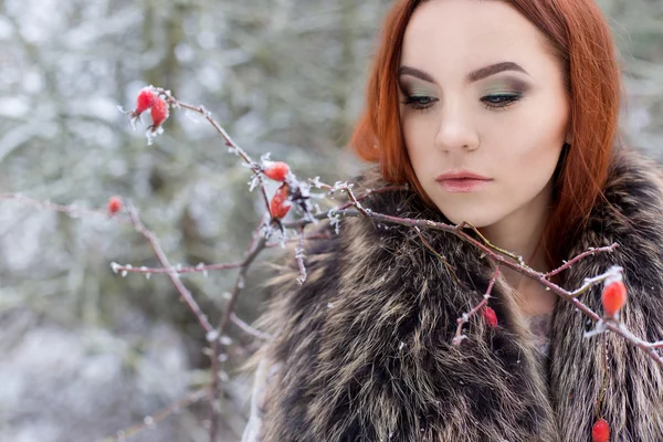 Beautiful cute sexy young girl with red hair walking in a snowy forest among the trees missed first trimester bushes with red yagodamiv warm coat with bright makeup