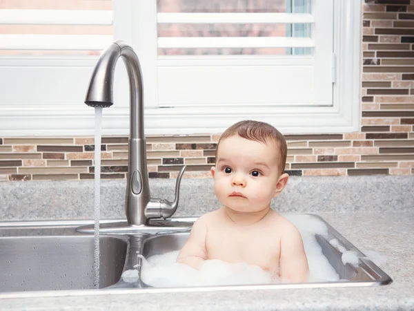 Portrait of cute Caucasian funny baby girl boy with dark black eyes sitting in big kitchen sink with water and foam  near window looking away, lifestyle everyday concept