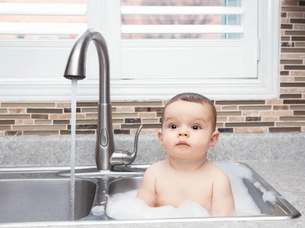 Portrait of cute Caucasian funny baby girl boy with dark black eyes sitting in big kitchen sink with water and foam  near window looking away, lifestyle everyday concept